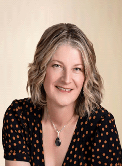 Smiling woman with wavy hair in a polka-dot dress, wearing a necklace, poses against a soft, neutral background, conveying warmth and approachability.