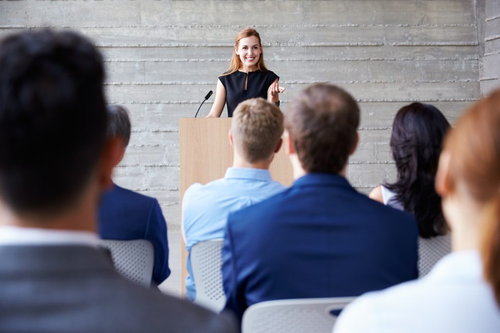 A speaker presents confidently at a podium, engaging an audience in a modern setting, emphasizing the importance of communication and presentation skills.