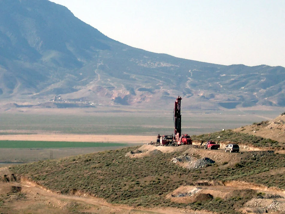 A drilling rig on a hillside with vehicles nearby, set against a mountainous landscape, highlighting resource extraction activities in the area.