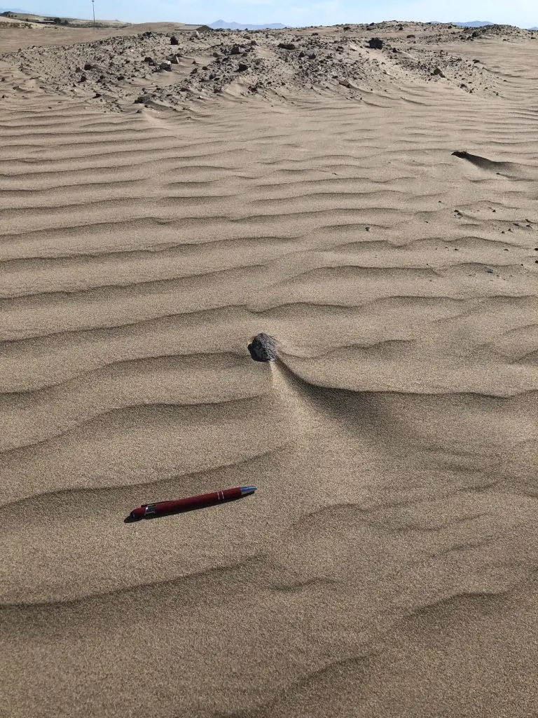 A maroon pen rests on textured sand dunes, showcasing the stark, barren landscape of a desert environment. The pen provides scale to the scene.